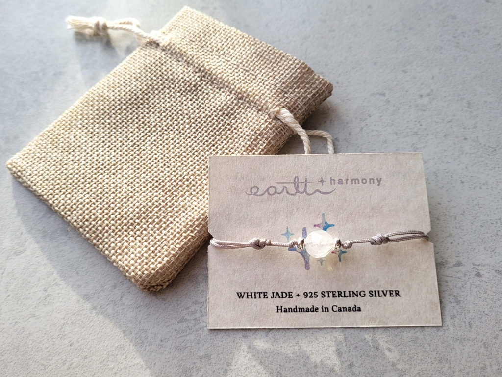 The bracelet (knotted cord style with one facet white jade, two 925 sterling silver beads, and magnetic clasp) is presented on a beige card with printed information. In the background is the jute bag the bracelet will come in.