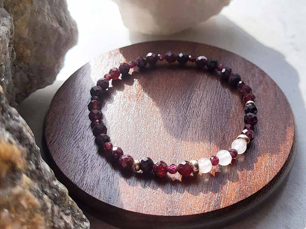 Garnet and white jade stretch bracelet is sitting on a dish for display. The sunlight is shining on it to show the detail in cut and also different sized beads. Garnet beads are facet cut in two sizes (5mm and 3mm), and features three round white jade beads (4mm) as central focus.